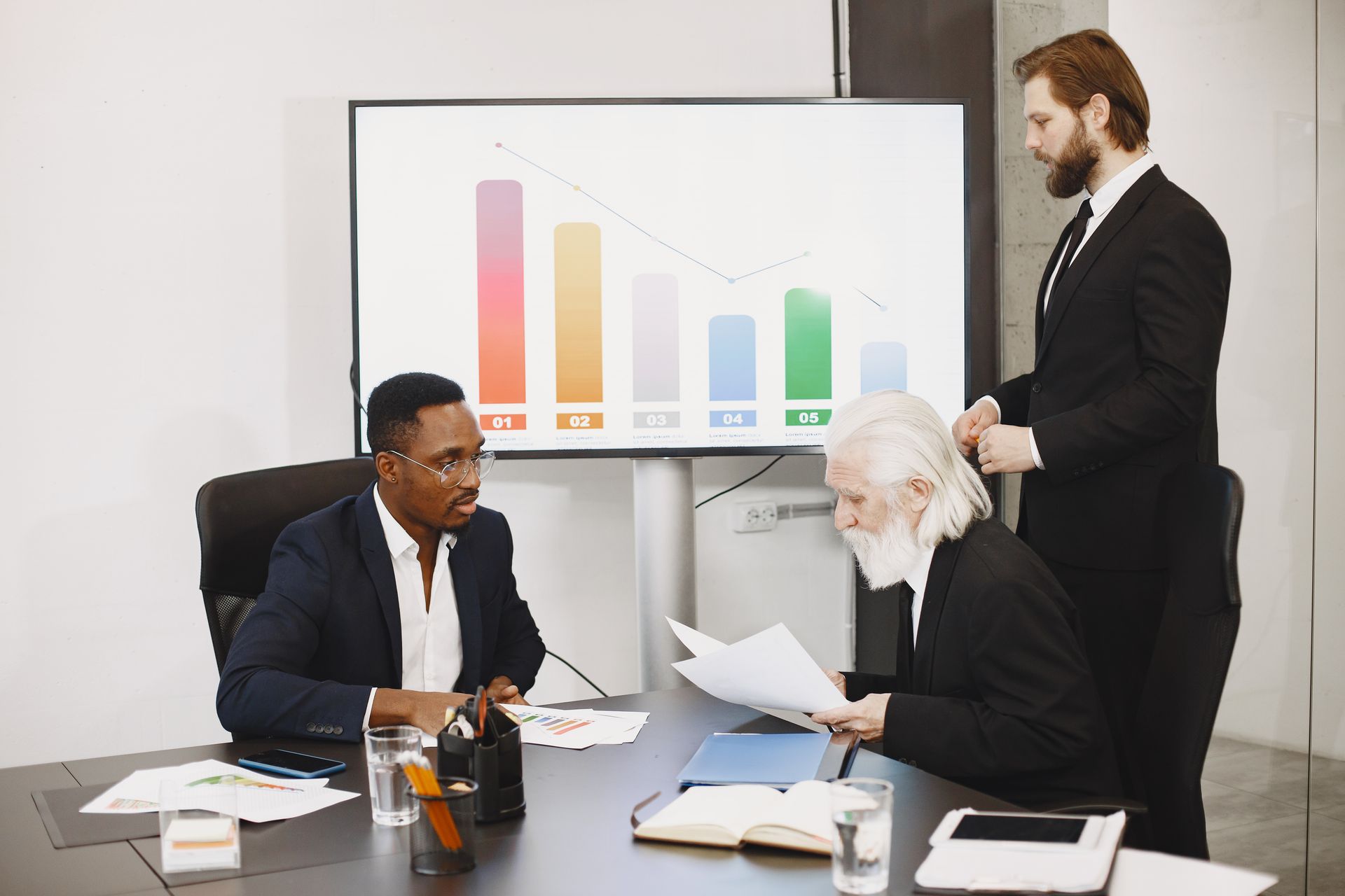 Three businessmen in dark suits at a boardroom table with a bar chart on the wall screen behind