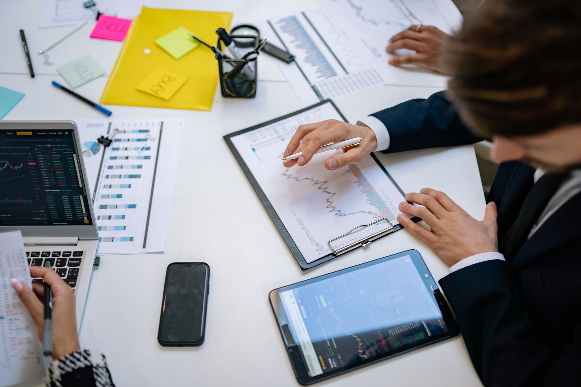 Overhead of a business desk with printed financial charts, a laptop showing a dashboard, tablets and hands sketching