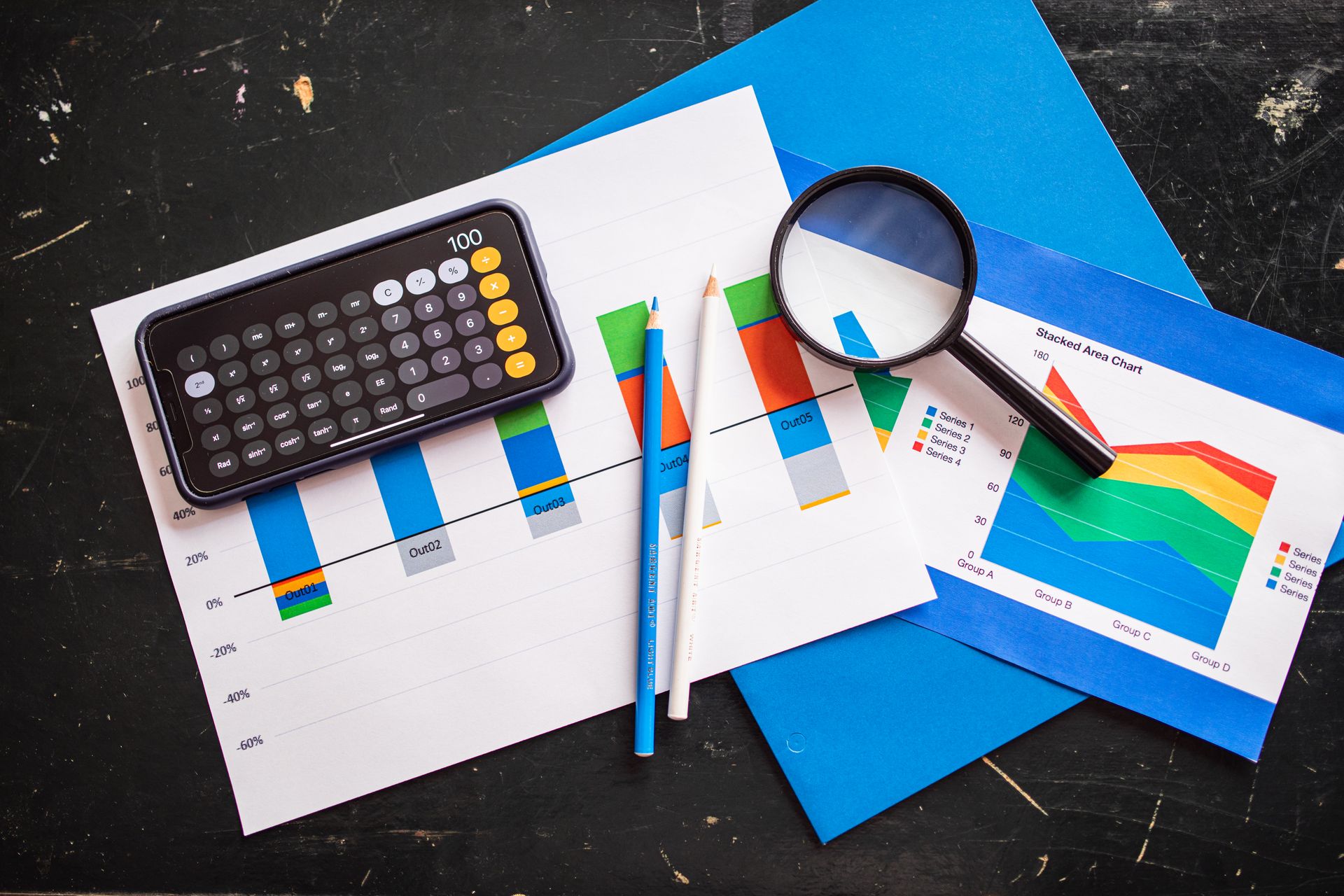 Overhead of colorful stacked area chart and blue bar graph with a magnifying glass on a dark desk